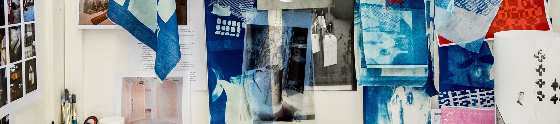 Colourful fabric samples attached to a noticeboard along with concept art; a pot of brushes, scissors and glue are visible at one corner of the board. 