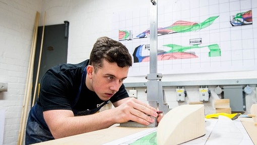 A young man stoops over a table, eyes fixed on a metal height gauge; the wall behind him displays plans for a sports car chassis.