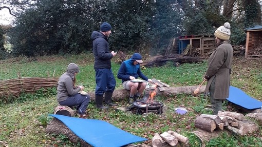 Two people sit on logs while two stand around a campfire; a big holly tree can be seen in the background.