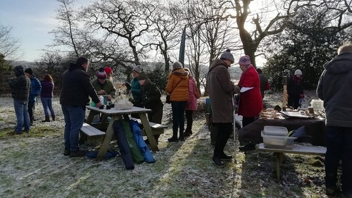 About fifteen people in thick winter jackets and hats stand around picnic tables in a frost-covered field.