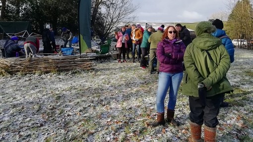People wrapped up against the cold stand talking in a wintry field; a few more take out supplies in an area enclosed by a wicker fence.