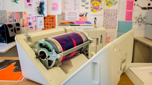 A colourful poster wrapped around a metal cylinder in a modern printing press. 