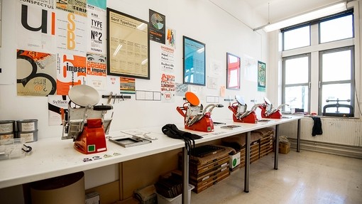 Four hand-operated red printing presses set out on a long workbench; the walls above them are decorated with typeface-themed posters.