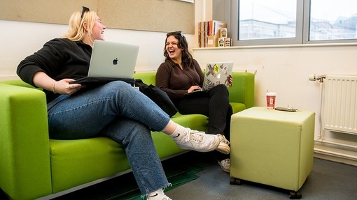 Two laughing students sit on a lime green sofa with MacBooks balanced on their laps.