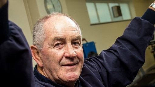 An older man looks towards the camera with his arms raised; a row of treadmillls stands behind him.