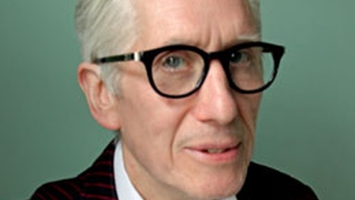 John Wood looks a little aside towards the camera through thick-rimmed sixties-style glasses; his white hair is brushed back from his face and he wears a pinstriped jacket suit.  