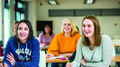        Myfyrwyr yn gwenu ac yn ysgrifennu ar lyfrau nodiadau mewn ystafell ddosbarth
