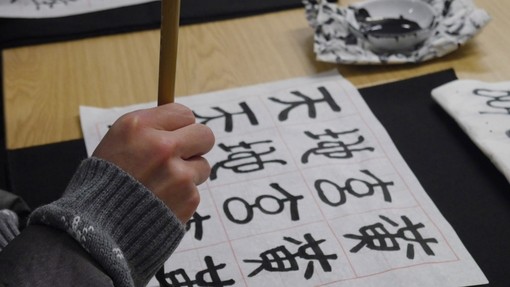 A hand holding a wooden calligraphy brush over a page of Chinese characters.