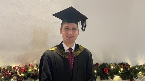 Brandon Gorman yn gwenu wrth wisgo het a gŵn academaidd sydd â chwfl  aur.