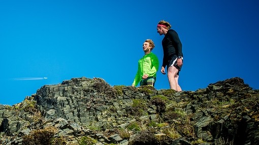 Two young men in running gear stand on an outcrop of rock below a bright blue sky.