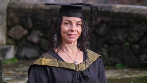 Mae Emma Williams yn gwenu fymryn wrth wisgo het a gŵn academaidd sydd â chwfl  aur.